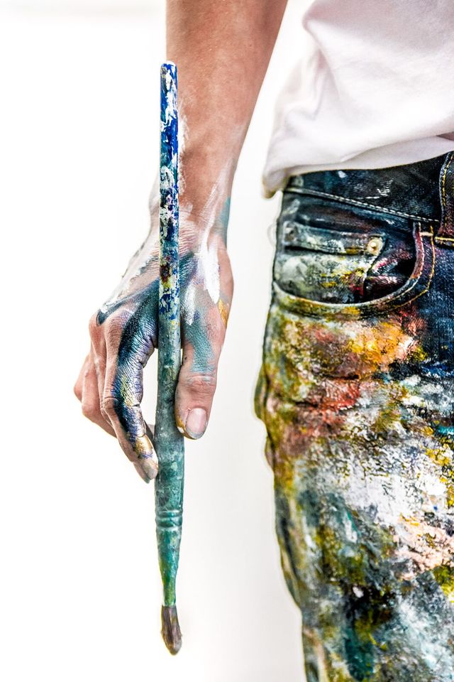 A photo of Donna Giraud in her studio in her paint covered jeans holding a paint brush.